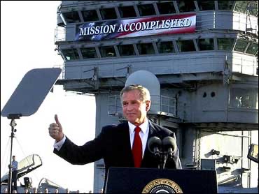 President George W. Bush standing at a podium in front of an aircraft carrier displaying a 'MISSION ACCOMPLISHED' banner, giving a thumbs up gesture.
