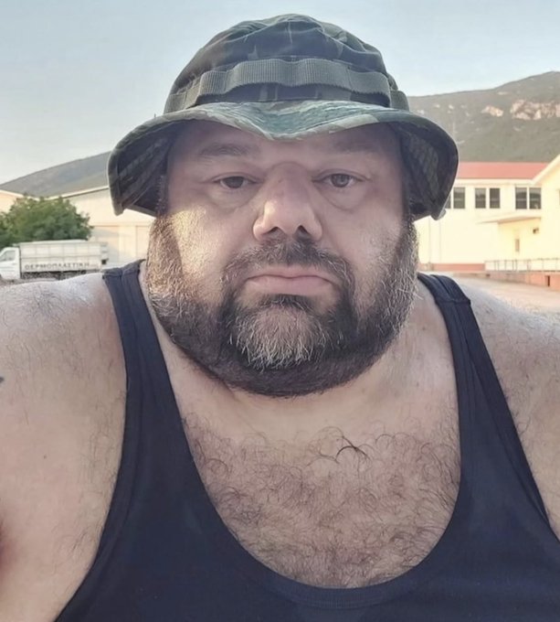 A selfie of a bearded man wearing a camouflage bucket hat and black tank top, standing outdoors with mountains and buildings in the background.
