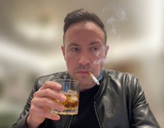 A man in a black leather jacket holding a glass of whiskey with a cigarette in his mouth.