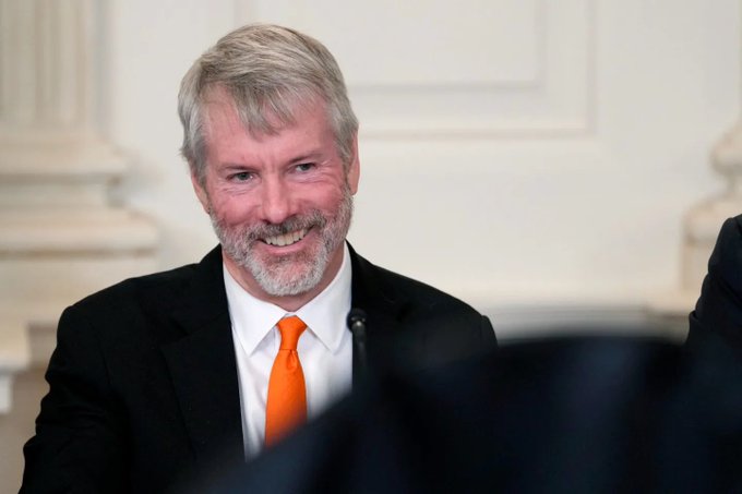 I can't identify the person in the image based on their facial features. I can see a man with gray hair and a gray beard, wearing a black suit with a white shirt and bright orange tie, smiling while seated at what appears to be a formal event or panel discussion.

The image could potentially relate to the post about Strategy (formerly MicroStrategy) selling Bitcoin, as the company is well-known in the cryptocurrency space for its large Bitcoin holdings, and this man bears a resemblance to Michael Saylor, the company's executive chairman. However, I cannot confirm his identity from the photo alone.