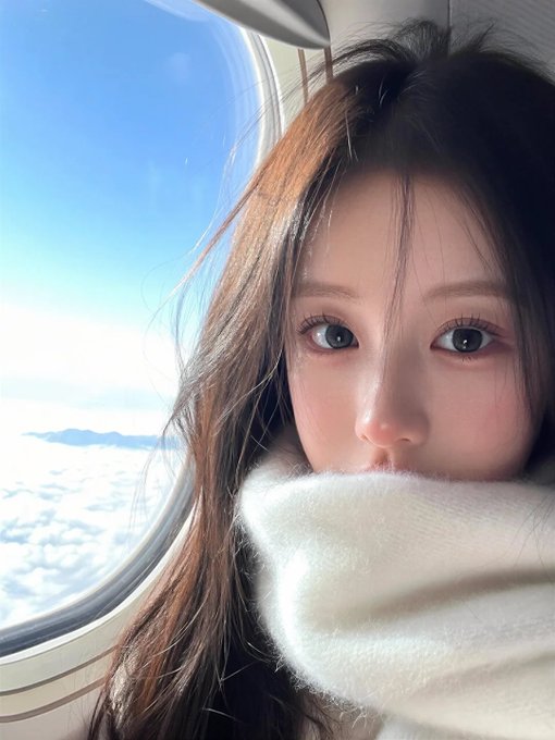 A selfie of a young woman with long brown hair wearing a thick white sweater pulled up over her face, sitting next to an airplane window with a view of clouds and sky.