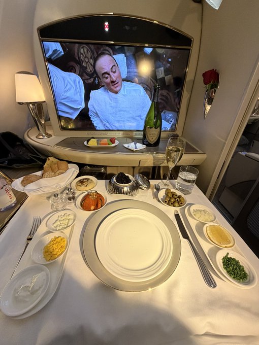 The image shows an Emirates business class in-flight dining setup featuring a private screen displaying a chef, a champagne flute, and an array of gourmet appetizers, caviar, and side dishes elegantly arranged around an empty central plate. The caption relates to the post by showcasing Emirates' premium luxury service, which travelers could soon book using cryptocurrency like Bitcoin through Crypto.com's payment solution.
