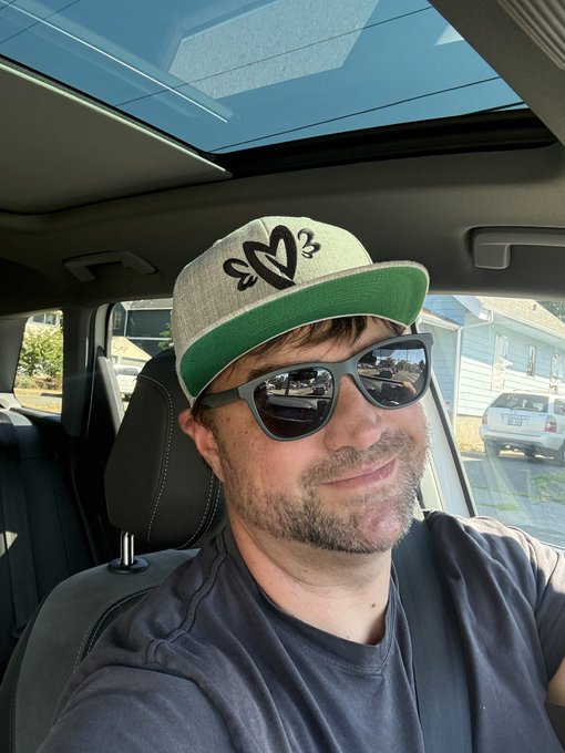 A man wearing a green and gray baseball cap with '2013' embroidered on it and dark sunglasses smiles while sitting inside a car with a sunroof.