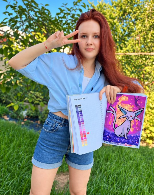 A red-haired young woman in a blue shirt and denim shorts posing with a peace sign, holding a folder with a Pokémon Espeon artwork and a color swatch palette.