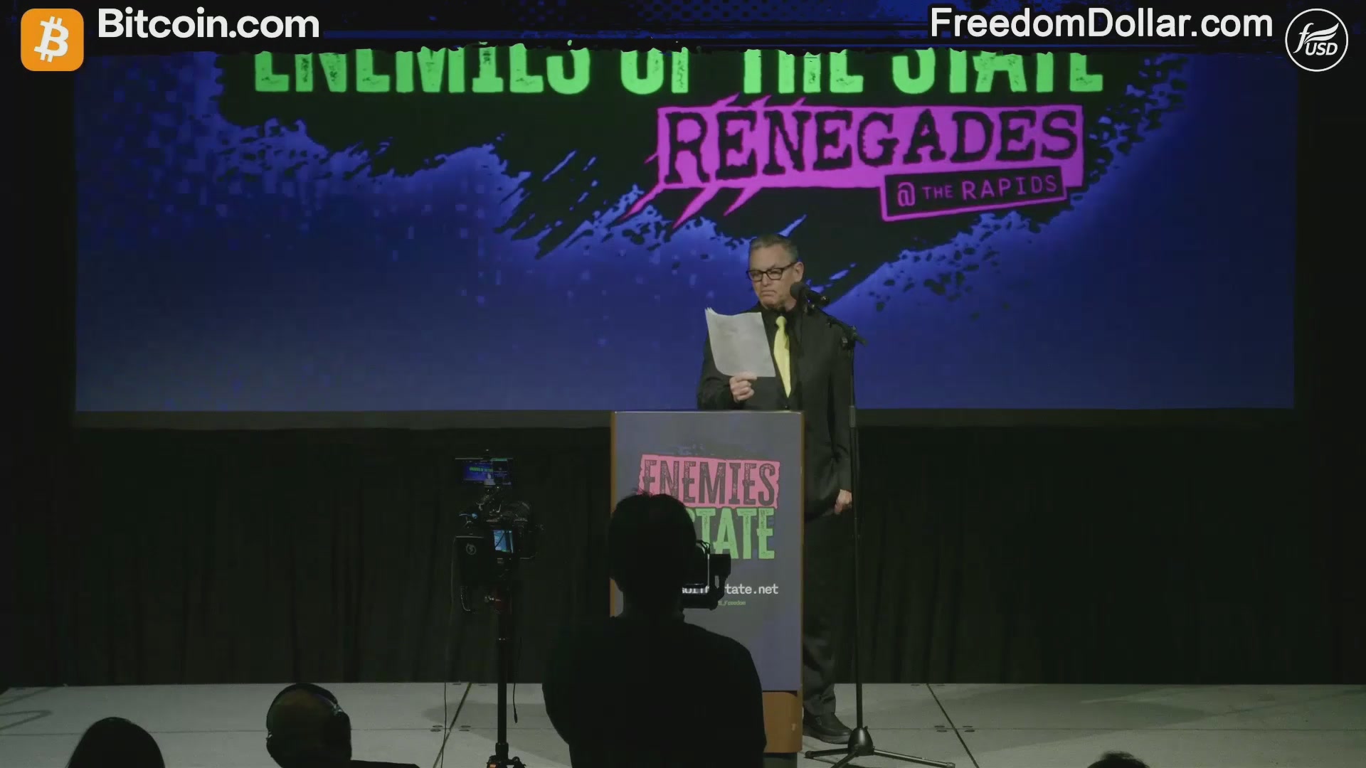 The image shows a man standing at a podium, giving a speech or presentation. The background of the image features a large screen displaying information about Bitcoin and its potential threats to privacy. There are two people visible in the scene - one person sitting behind the podium and another person sitting on the side of the stage.