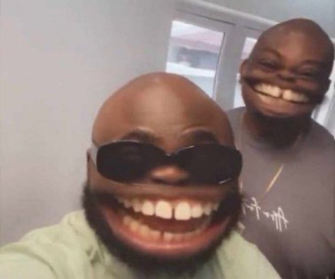 A meme image featuring two smiling Black men in an indoor setting, with the man in front wearing sunglasses and a green shirt, and the man behind wearing a brown shirt.
