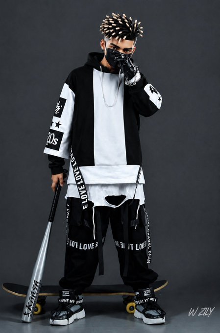 A full-body studio portrait of a man with dreadlocks wearing a black face mask, a black and white hoodie with 'E LOVE' and '20s' text, black pants, and holding a baseball bat while standing on a skateboard.