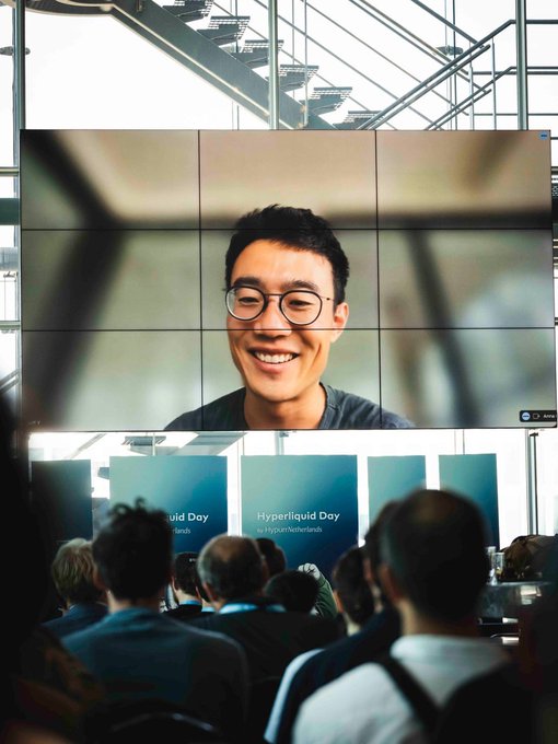 An audience views a large screen displaying a smiling man with glasses; banners below read 'Hyperliquid Day by Hyperliquid Labs.'