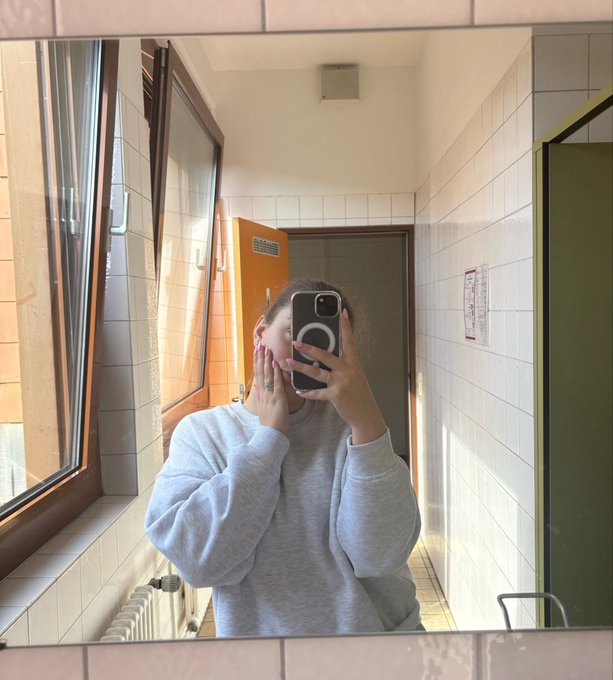 A mirror selfie of a person in a gray sweatshirt holding a phone with a circular grip, standing in a tiled hallway with windows on the left side.
