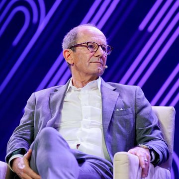 The main focus of this image is a man sitting down with a thoughtful expression while wearing glasses. There are other people visible in the scene, but they don't seem significant to the main subject. The background features purple lines, which create an artistic and intriguing backdrop for the image. Additionally, there appears to be some text that reads "BREAKING Bitcoin miner MARA appears to have acquired 1,000 BTC worth roughly $66.7M", implying that the man sitting down might be associated with or related to a significant event involving cryptocurrency.