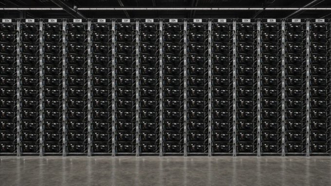 A photograph of a large server rack room filled with rows of black mining equipment or servers with illuminated indicators, set in an industrial facility with concrete flooring.