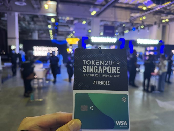 A hand holding a Token2049 Singapore attendee badge and Visa Platinum card at Marina Bay Sands, dated 7-8 October 2026, with a blurred event venue in the background.