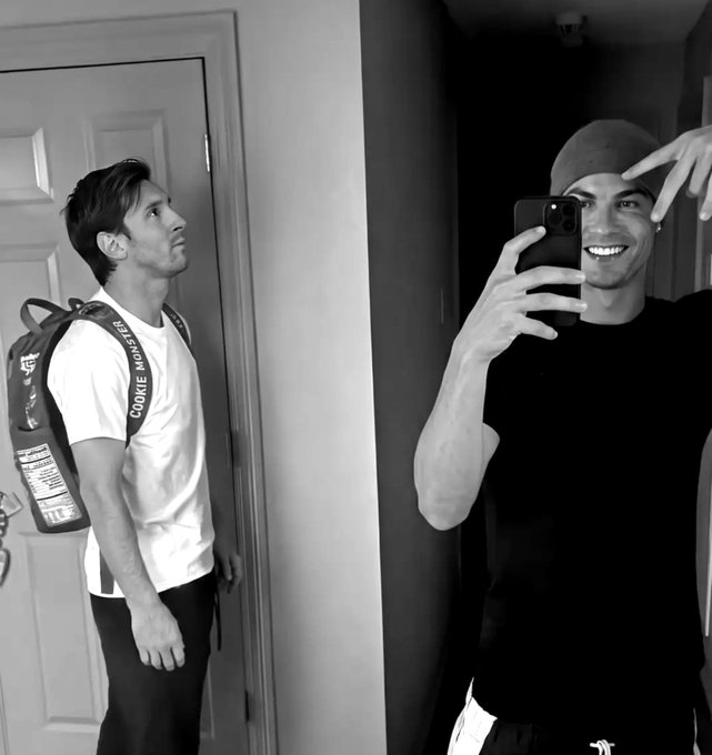 A black-and-white mirror selfie of Cristiano Ronaldo smiling while holding his phone, with Lionel Messi standing behind him wearing a backpack and looking away.