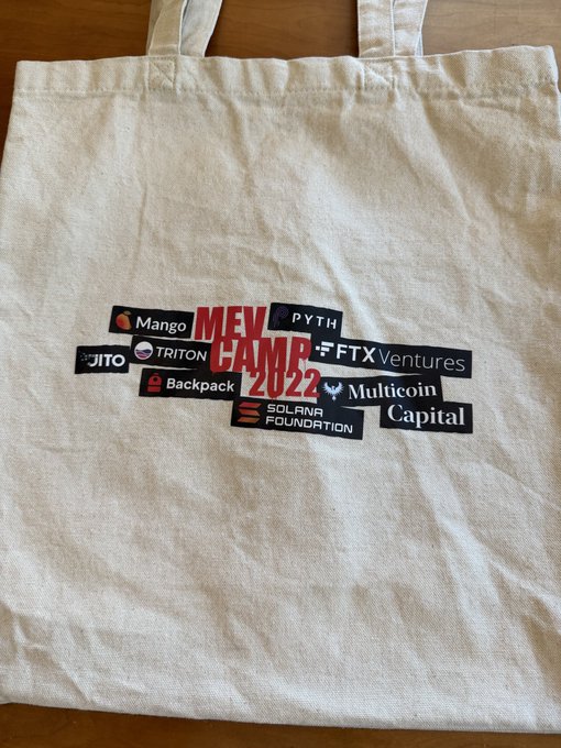 A cream-colored tote bag from MEV Camp 2022 rests on a wooden surface, displaying sponsor logos including Mango, Pyth, Triton, Jito, FTX Ventures, Backpack, Multicoin Capital, and the Solana Foundation. The image illustrates the speaker's reminiscence about their early presentation on Solana's Geyser streaming interface, capturing a piece of memorabilia from a pivotal moment in MEV (Maximal Extractable Value) and Solana DeFi history.
