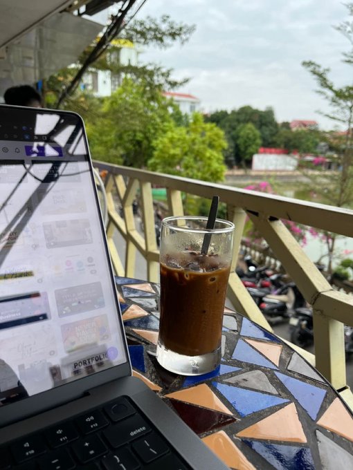 A glass of iced coffee sits on a colorful mosaic tile table next to a laptop, with a street scene of motorbikes and greenery visible beyond a railing.