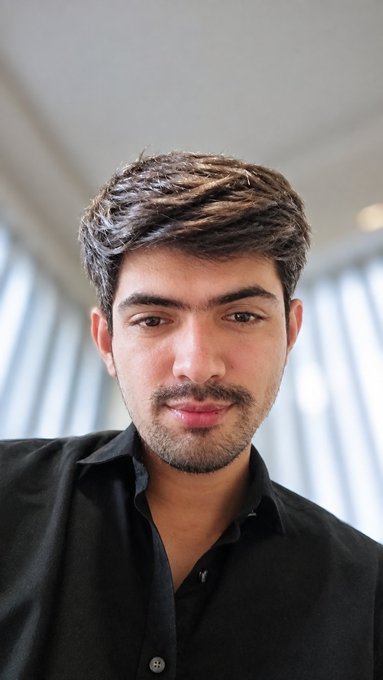 A young man with dark hair and a short beard looks at the camera while wearing a black button-up shirt, with bright vertical blinds in the background.