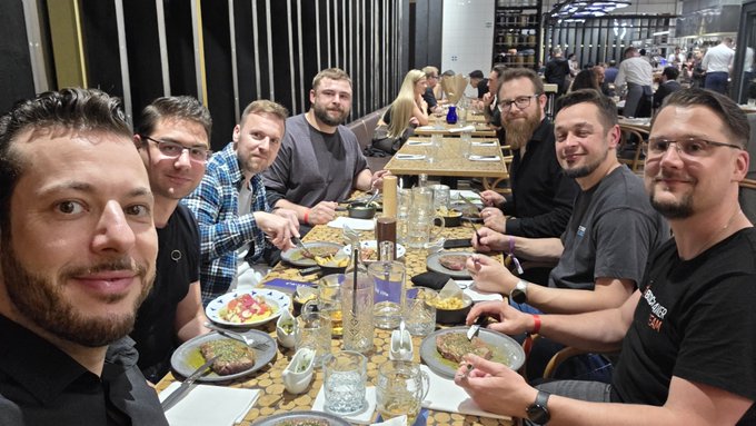 A group of eight men gather around a long table enjoying a team breakfast together at what appears to be a restaurant, with plates of food, glasses, and bottles spread across the table. The image captures a friendly team camaraderie moment before the #BlocktrainerEvent, as mentioned in the accompanying post.