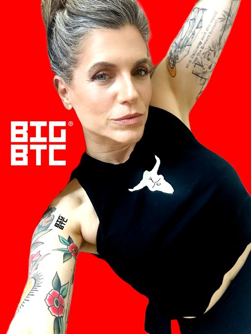 A woman with short gray hair, a tattooed arm, and a black tank top poses against a vibrant red background featuring the "BTC BTC" logo with a registered trademark symbol, capturing the bold and rebellious spirit of the "#GM®" rallying cry in the crypto community.