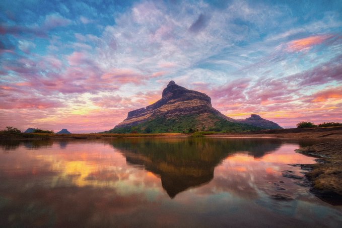 A stunning landscape featuring a large mountain reflected in a calm body of water at sunset, with pink and blue clouds filling the sky.