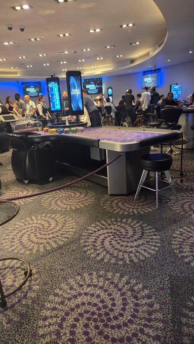 A bustling casino floor at 3 AM with patrons crowded around electronic gaming tables and slot machines, illuminated by glowing digital screens under recessed ceiling lights, perfectly capturing the late-night gamble of staying awake to launch on Pump.fun instead of sleeping. Visible signage includes "Double Black Diamond" and "Peach & Win" displays reinforcing the all-night, high-stakes atmosphere.