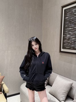 A young woman wearing sunglasses, a black hoodie, and black shorts poses indoors next to a beige sofa.