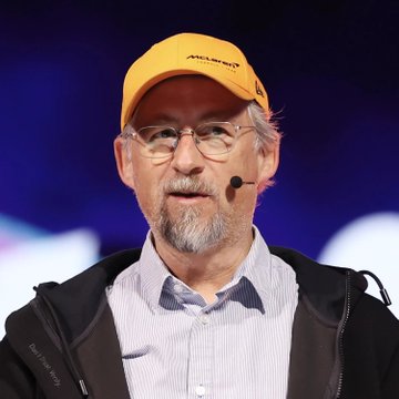 A close-up photo of an older man with a gray beard wearing a yellow McLaren cap, glasses, and a black jacket over a light blue shirt.