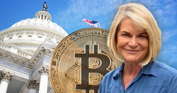 A blonde woman, identified by the accompanying post as Senator Cynthia Lummis, smiles while wearing a blue denim shirt, positioned in front of a composite background featuring the U.S. Capitol dome and an oversized gold Bitcoin coin inscribed with "BITCOIN DIGITAL DECENTRALIZED PEER TO PEER." The imagery visually connects her urgent push for the CLARITY Act to the cryptocurrency and legislative worlds, symbolizing her advocacy for clear digital asset regulations.