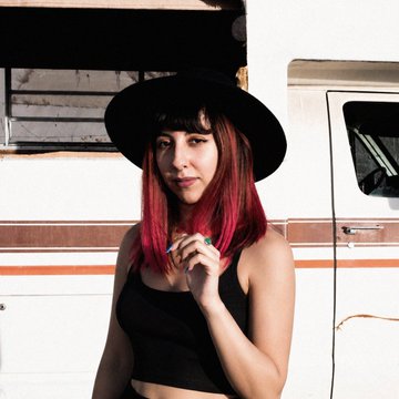 A portrait of a young woman with red hair wearing a black wide-brimmed hat and a black top, standing in front of a white van or RV.