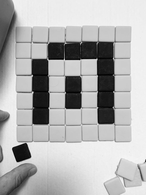 A black-and-white photograph of hands arranging black and white square tiles into a pixel-art pattern resembling a pixelated letter or symbol on a grid.