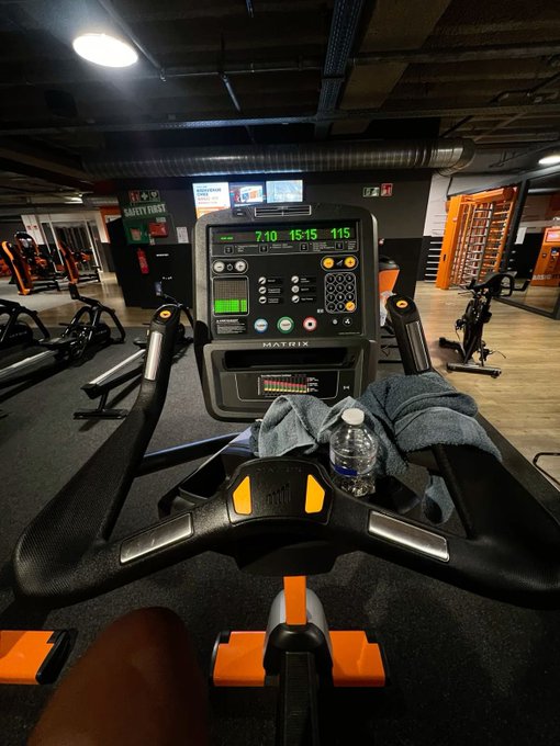 A first-person view from a Matrix-brand stationary bike in a dimly lit gym, with the console displaying metrics like "7:10," "15:15," and "115," alongside a water bottle and towel resting on the handlebars. The post's "GM CT" (good morning Connecticut) and "Happy taco Tuesday" tie into a morning fitness session before enjoying tacos later.