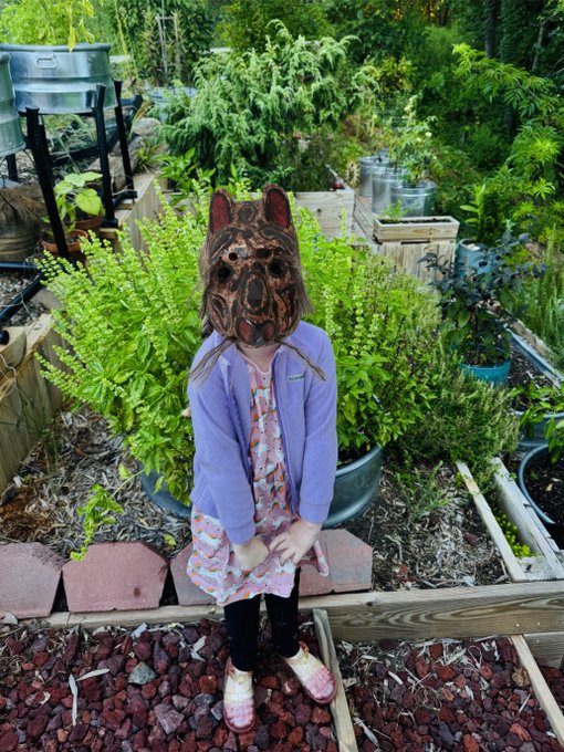 A young child stands in a lush garden wearing a floral dress, purple cardigan, and a carved wooden animal mask, posed in front of raised wooden garden beds.