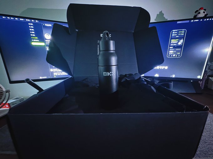 A black OKK branded water bottle displayed inside its opened presentation box on a desk, with dual computer monitors visible in the background.