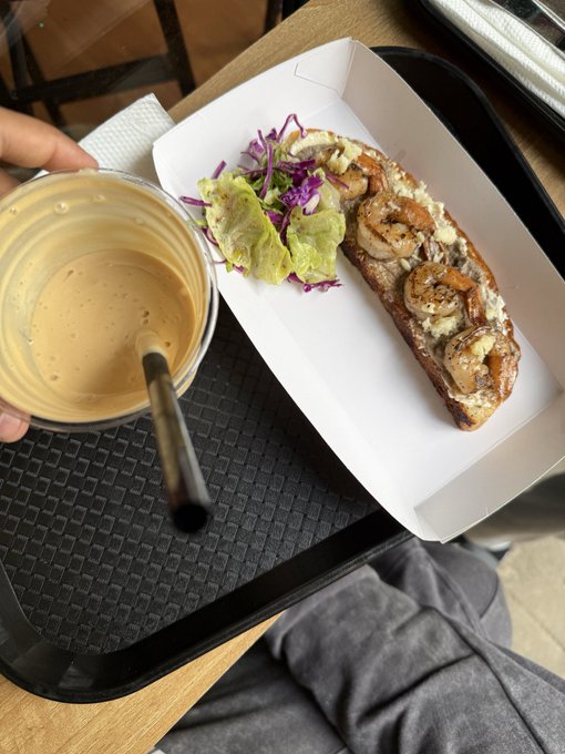 A tray holding a plastic cup with a creamy orange drink and a straw, alongside a sandwich or open-faced bread topped with shrimp and guacamole in a takeout container.