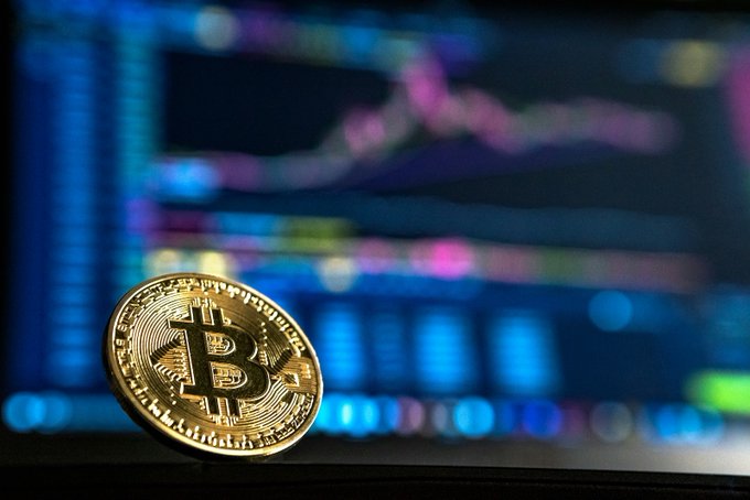 The image features a close-up view of a gold-colored Bitcoin coin sitting on top of a computer monitor or laptop screen, positioned at the bottom center portion of the frame. In the background, there's a computer screen displaying financial data that seems out of focus. The post related to this image talks about Bitcoin's historical price behavior and its current state. It highlights how Bitcoin's value has been falling, with over half of the Bitcoin supply now underwater, suggesting it is approaching or below a final leg in a market cycle.