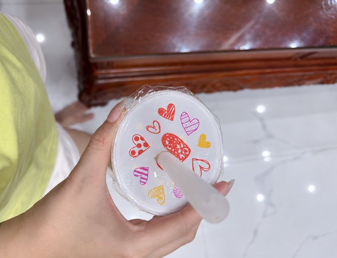 A hand holds a small round container decorated with colorful heart designs wrapped in plastic, sitting on a marble floor near a wooden furniture piece.