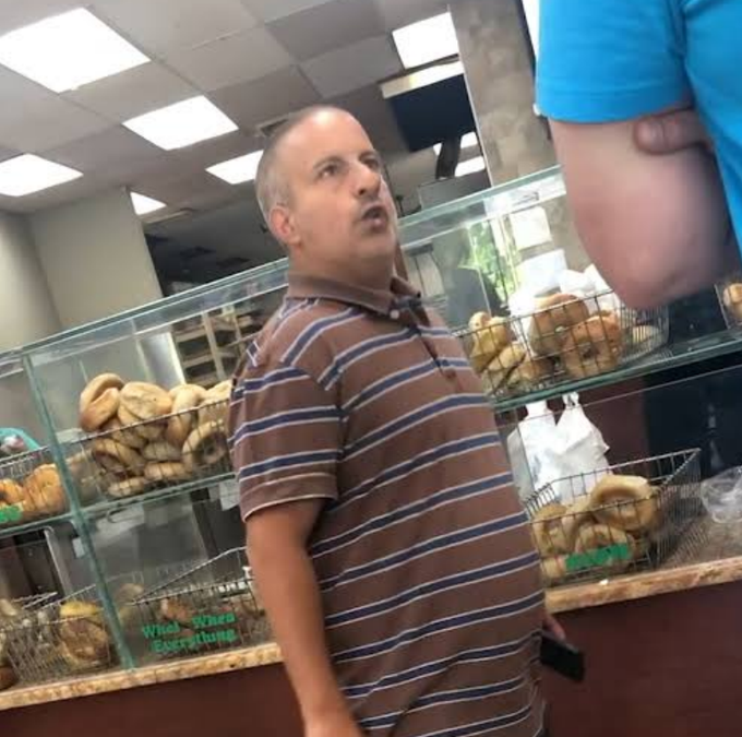 A balding man in a brown striped polo shirt stands in a bagel shop next to a glass display case filled with bagels, gesturing while speaking, with a sign reading 'What...When... Everything' visible on the case.