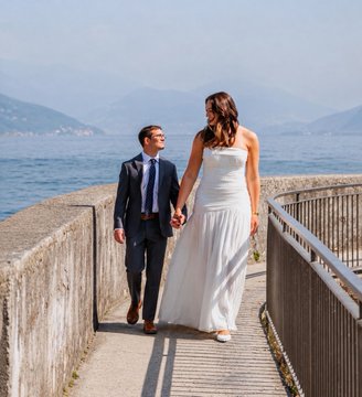 A bride and groom walk hand-in-hand along a waterfront promenade, dressed in formal wedding attire with a serene lake and mountains in the background. The post "So proud of my boy @marccolcer" suggests a parent is sharing this touching moment of a loved one's special day.