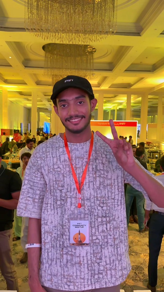 A young man wearing a black Bybit cap and a lanyard that reads "BYBIT COMMUNITY MEETUP" poses with a peace sign at the Bybit Community Meetup event in Lahore, set in an elegant venue with ornate chandeliers. His event badge features a small design referencing what appears to be the Eiffel Tower.