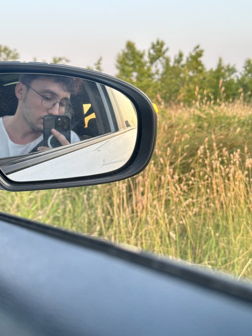 A side mirror of a car reflects a young man with glasses taking a selfie with his phone, set against a backdrop of tall grass and trees.