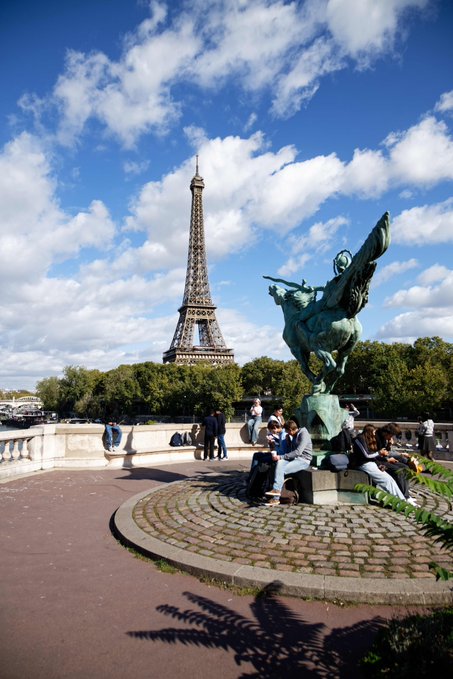 A scenic view of the Eiffel Tower in Paris seen from across the Seine, with a bronze equestrian statue in the foreground and tourists seated on benches under a partly cloudy blue sky.