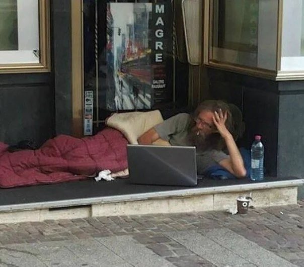 The image shows a man lying down on the ground, using his laptop computer while surrounded by various objects such as a bottle of water, a cup, and some trash nearby. The scene appears to be set outdoors with a brick sidewalk and a building in the background. The photo also contains text that reads "Me buying another BTC dip:". This suggests that the man might be involved in an online transaction or has recently purchased Bitcoin, which is often associated with digital currency transactions.