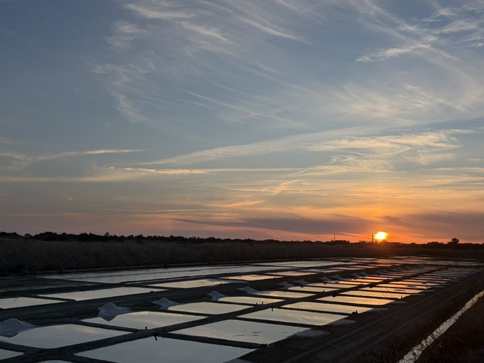 A scenic sunset photo over a landscape of salt flats or shallow pools, with the sun dipping below the horizon and wispy clouds in the sky.