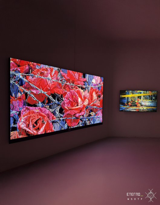 An installation view showing two digital screens mounted on a purple wall in a gallery space; the larger screen displays a close-up of red roses entangled in barbed wire, while the smaller screen shows a ship at a dock, with a logo for 'ETERNO MSOTO' in the corner.