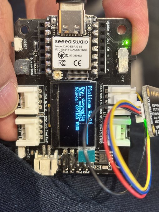 The image shows a person holding up a small electronic device that appears to be a circuit board or a computer chip with a display screen. The device has a green light, indicating that it is powered on. The device seems to be used for building hardware wallets, as suggested by the text in the image. The person's hand is visible near the bottom of the frame, and they are wearing a black shirt.