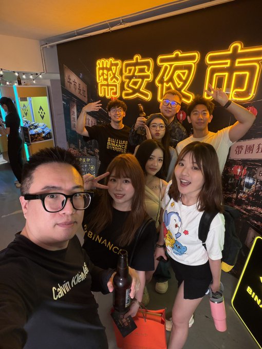 A group selfie taken at a Binance-branded event with a neon yellow sign reading '币安夜市' (Binance Night Market) in the background, showing several people smiling and posing together.