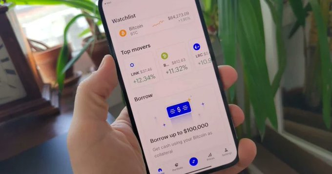 A close-up of a hand holding a smartphone displaying a crypto trading app, showing a 'Watchlist' with Bitcoin ($84,273.09, +1.00%) and 'Top movers' including LINK and LBC, along with a 'Borrow up to $100,000' option.