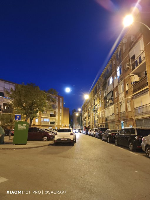 The image shows a street scene at night with several parked cars lining both sides of the road, including some black ones. The sky above appears dark blue, creating an atmosphere of nighttime tranquility. A green trash can stands out against the darkness on one side of the street. In the background, there are buildings that add depth to the scene and provide context for the location.