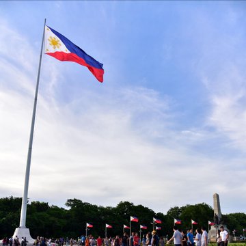 The Philippine flag waves prominently on a tall flagpole against a blue sky, with crowds and other flags visible at what appears to be a national monument. The image symbolizes the Philippines, the country where the Bank of the Philippine Islands is piloting stablecoin cross-border payment solutions for freelancers and remote workers.