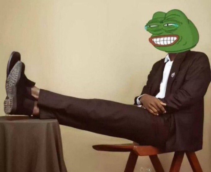 A photo of a man in a black suit with his feet up on a table, his head replaced with a smiling, crying green Pepe the Frog meme face.