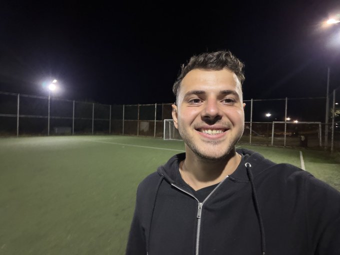 A selfie of a smiling young man in a black hoodie taken at night on an illuminated artificial turf soccer field with stadium lights and a goal visible behind him.